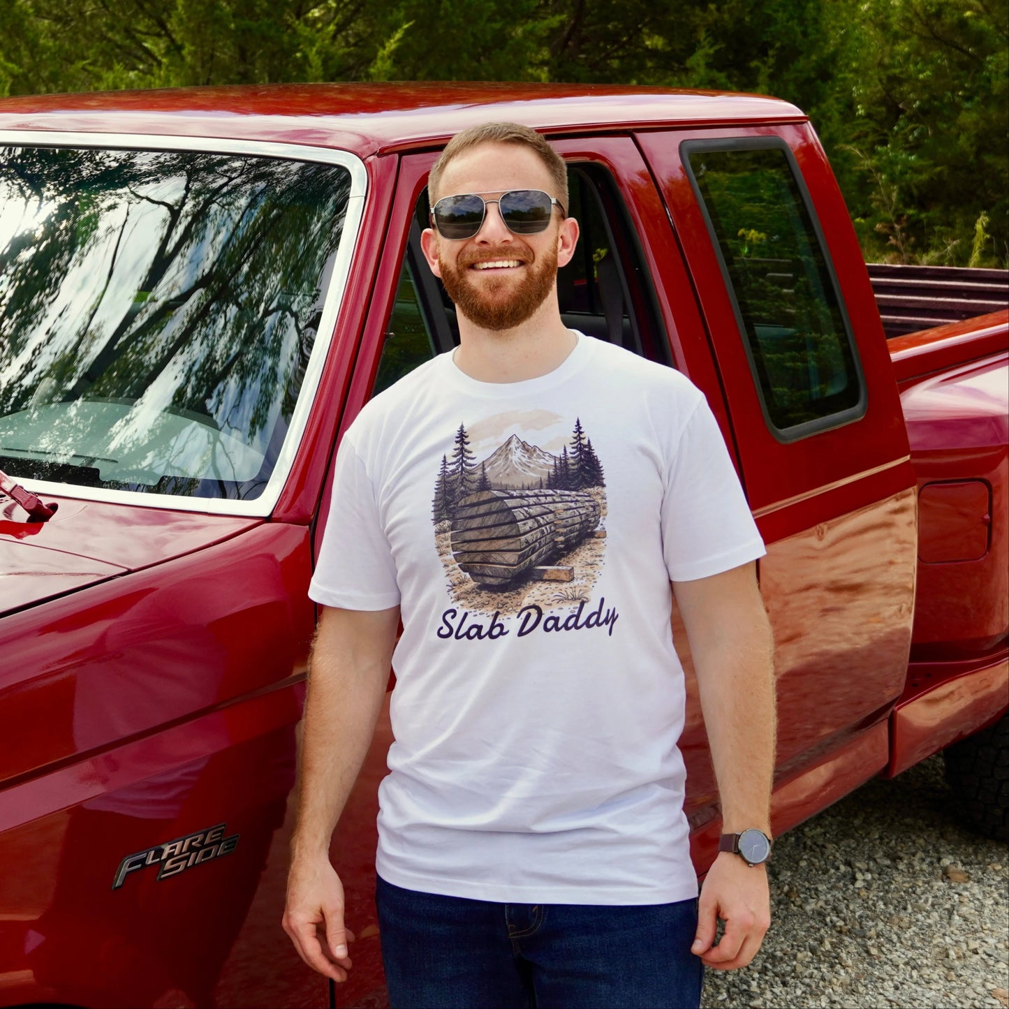 Man standing next to a red pickup truck with trees in the background