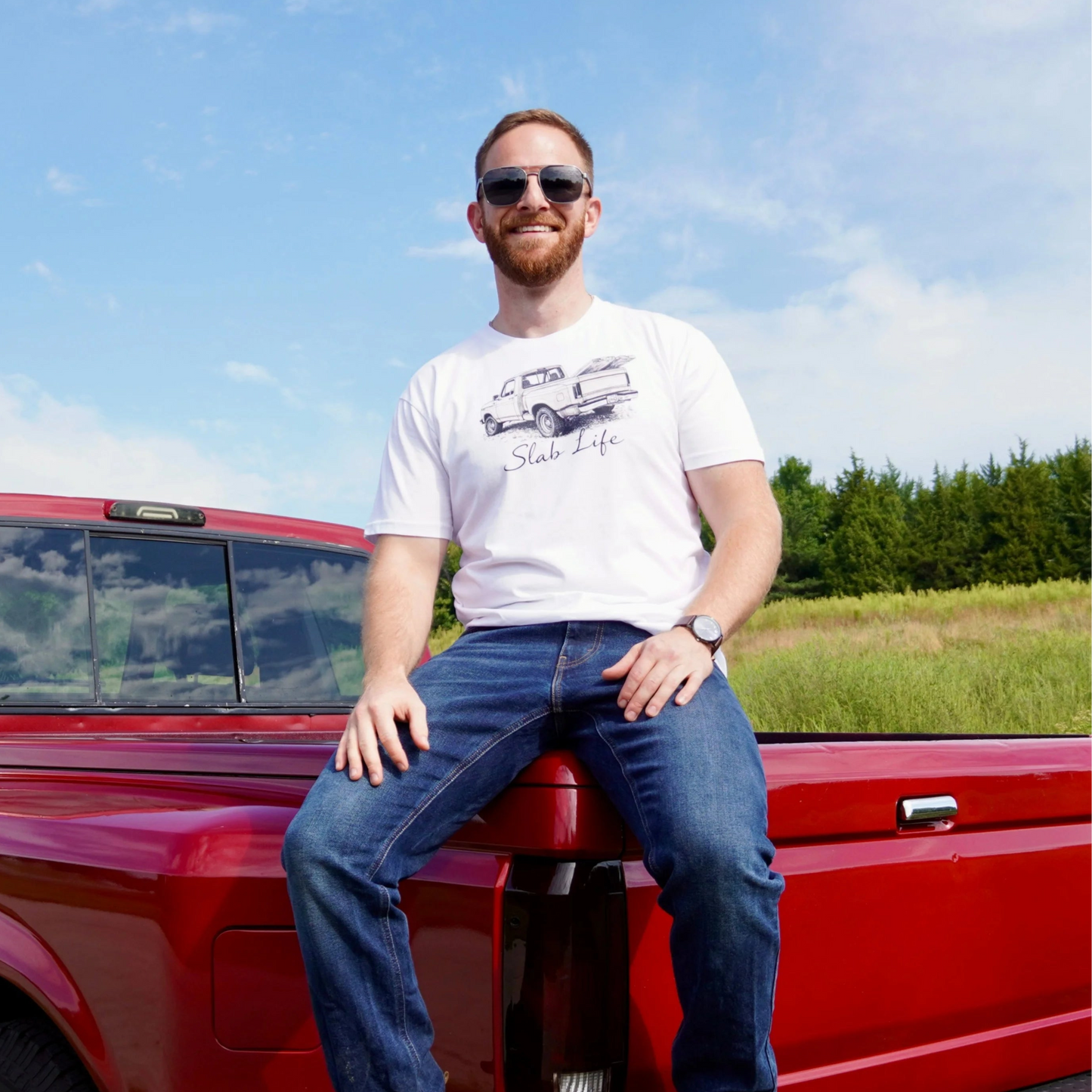 Man sitting on a red pickup truck in a field with trees in the background