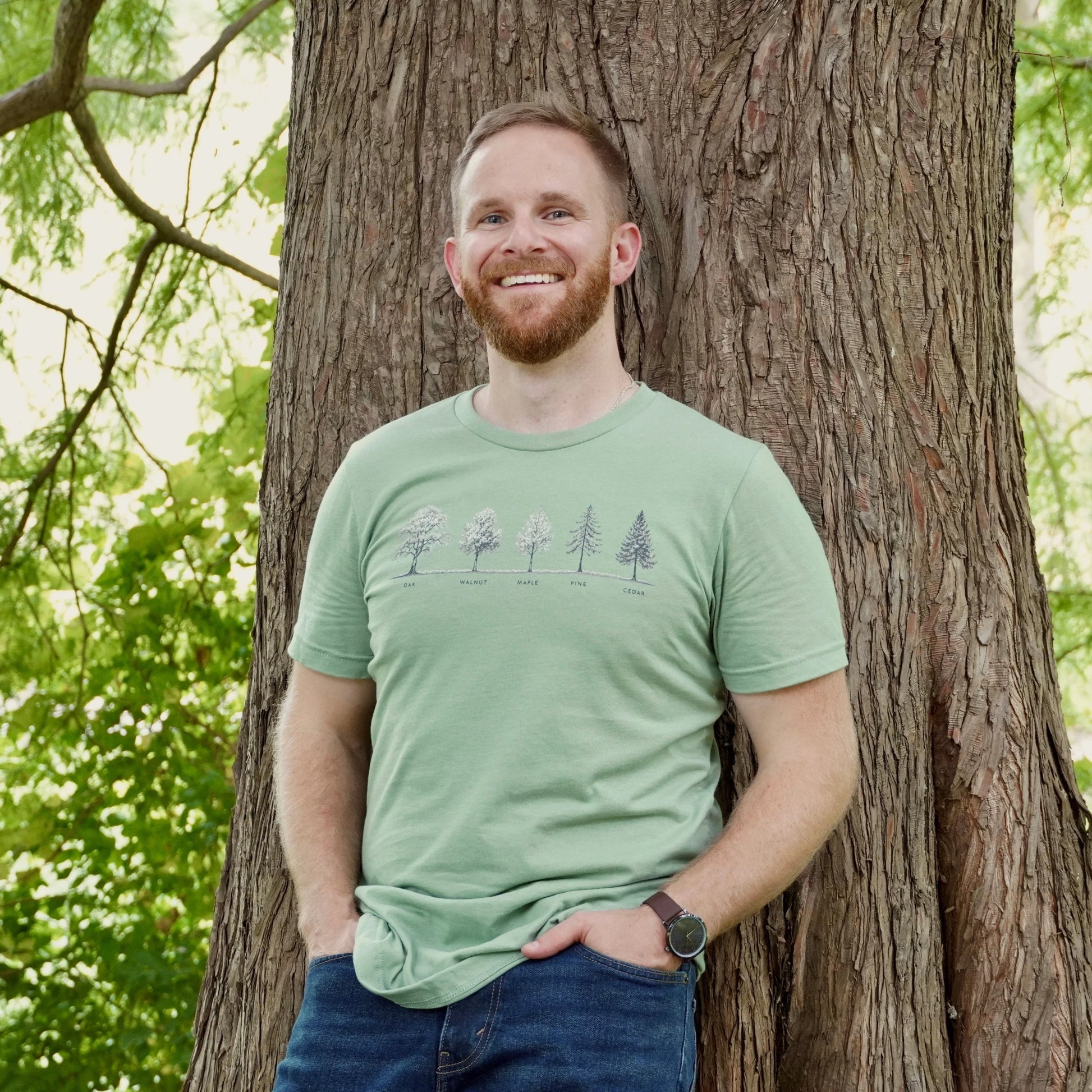 Man wearing a green t-shirt with a tree design, standing next to a tree in a forest setting.