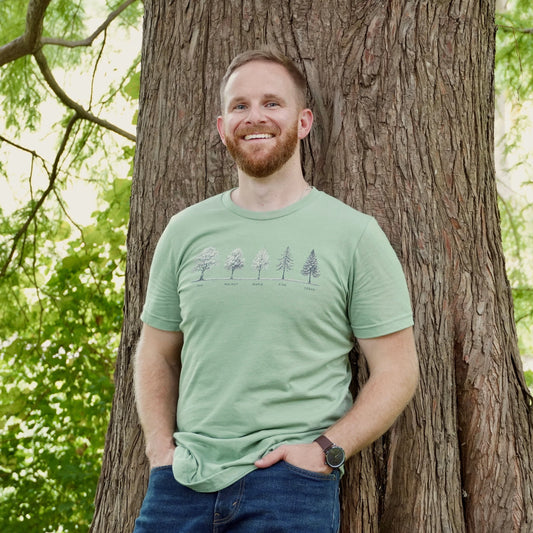 Man wearing a green t-shirt with a tree design, standing next to a tree in a forest setting.